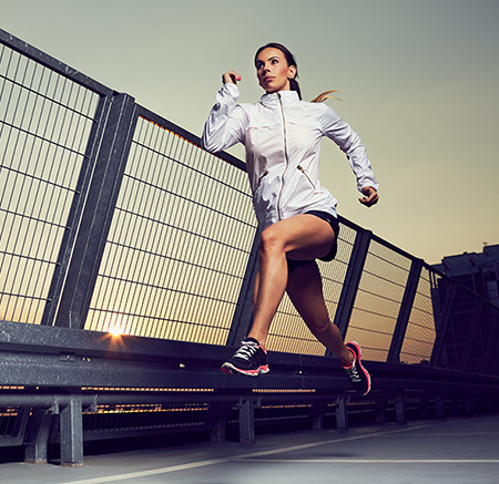 A woman in athletic attire is captured mid-run on a bridge with a fence, under an overcast sky during twilight hours.