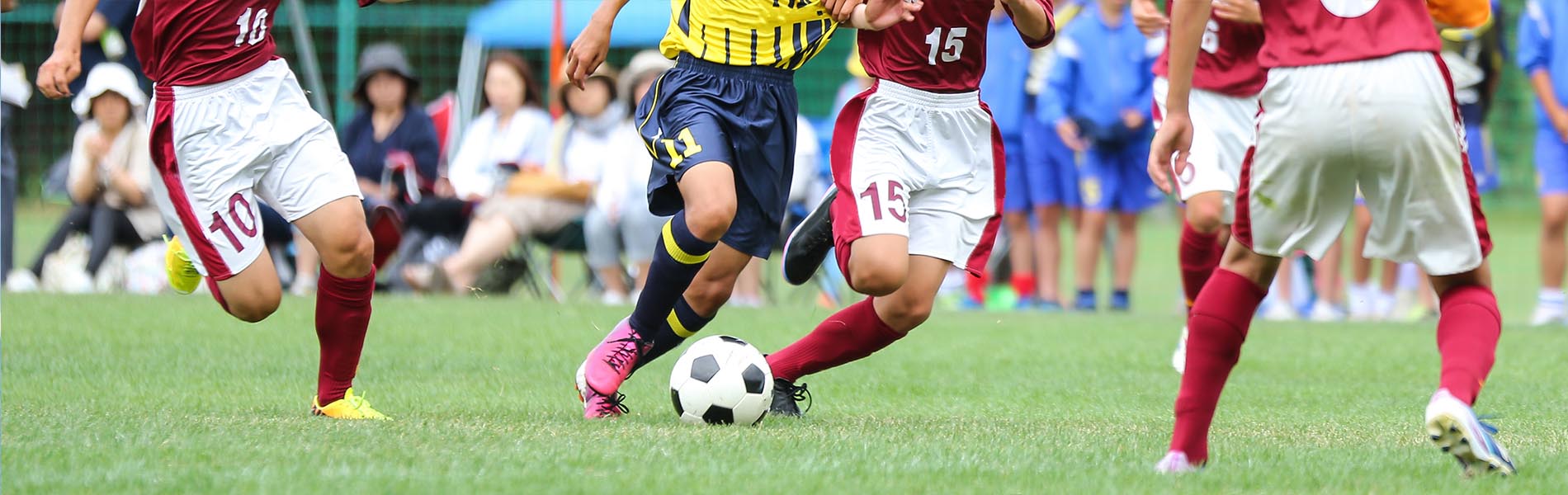 The image depicts a group of male soccer players on a field wearing uniforms with red and white colors, while one player wears blue, engaged in a game, with one player in focus kicking the ball towards a goal, and others in various states of motion around him.