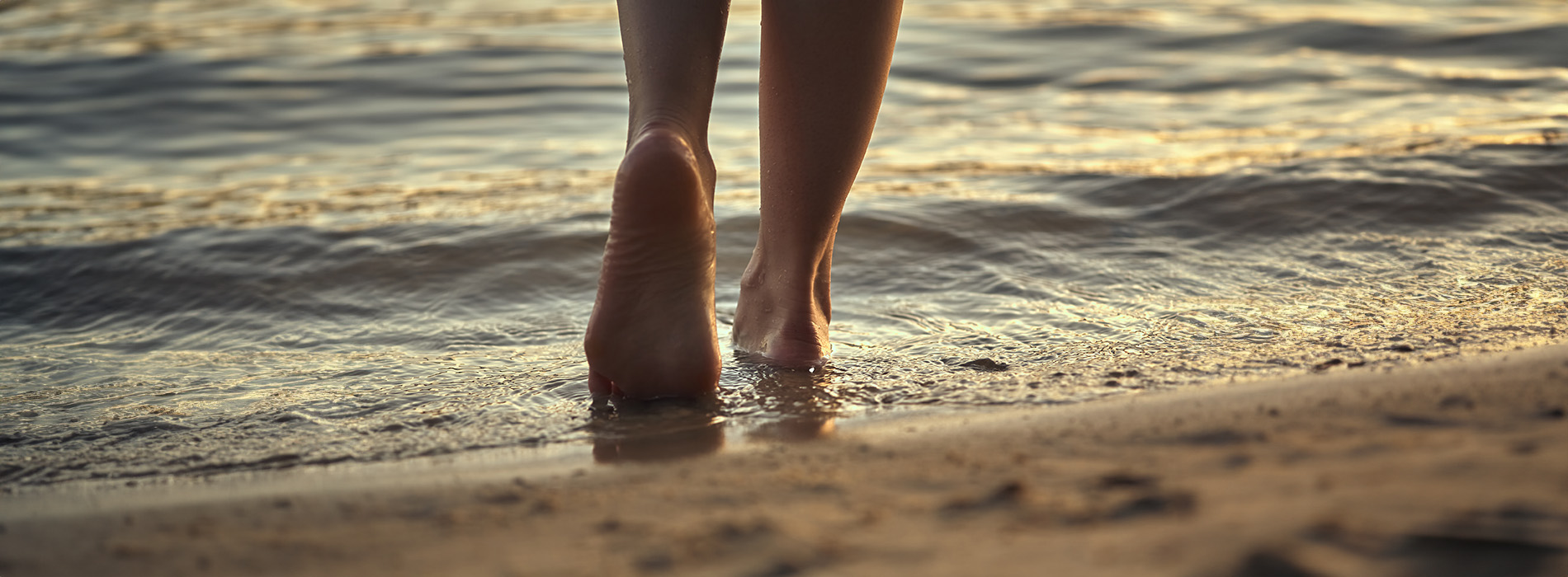 The image shows a person walking along the shoreline at dusk, with their feet submerged in shallow water, looking out towards the horizon.