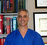 The image shows a man standing in front of bookshelves with framed certificates behind him, wearing a blue surgical scrubs top and a white stethoscope around his neck, smiling at the camera.